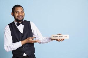 Hotel concierge guiding guests with a wall sign to find all amenities, presenting indicator with the direction to the restaurant against blue background. Confident skilled reception personnel. photo