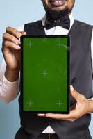 Bellman hotelier showing a tablet with green screen display in studio, presenting mockup chroma key display against blue background. Young hotel concierge working at a resort. photo