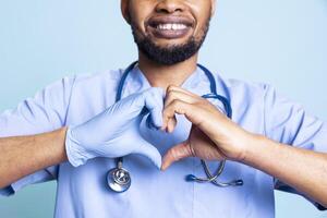 Portrait of smiling jolly specialist presenting heart shape sign with rubber glove on a hand. Expert nurse showing gratitude and love gesture on duty, standing against blue background. photo