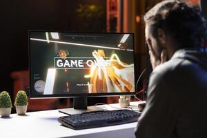 Player upset after losing while playing first person shooter videogame, putting face in hands in frustration. Man feeling dejected after receiving game over screen while playing on PC, crying photo