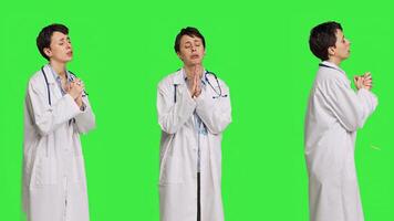 Hopeful physician holding hands in a prayer sign against greenscreen backdrop, asking for peace and forgiveness in studio. Woman medic specialist in white coat does meditation. Camera B. photo