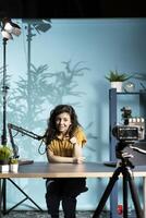Jolly woman filming vlog pointing finger to high tech camera in professional studio, speaking into microphone equipment. Smiling influencer addressing viewers , talking with them photo