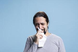 Sad melancholic person with anxiety crying and whining on camera, feeling distraught and in an emotional crisis against blue background. Young guy wiping his tears with a tissue. photo