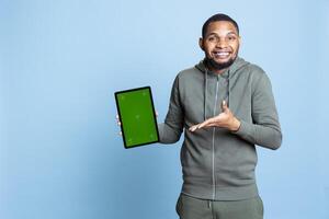 African american person in casual clothes shows mockup screen on tablet, pointing at the display and smiling for the camera, Young adult feeling happy and doing marketing content. photo