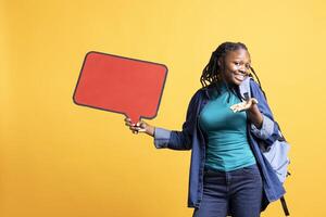 Cheerful woman using red speech bubble sign with empty copy space as advertisement. Upbeat student with backpack presenting thought bubble cardboard used as promotion concept, studio background photo