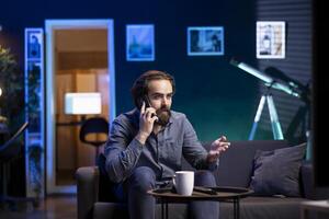 Person on couch binging series on streaming service, talking on mobile phone with friend. Relaxed man watching shows on television, discussing them with mate during telephone call photo