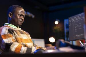 African american self employed developer in home office coding on his computer at workstation. Focused IT specialist doing contracted job from apartment, writing lines of code at desk. photo