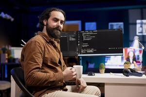 Portrait of cheerful computer engineer enjoying hot beverage while implementing security measures to protect company systems and data. Smiling IT worker drinking coffee and debugging code photo