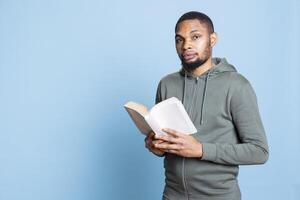 African american cheerful guy enjoys reading a book in studio, feeling happy with literature and novel against blue background. Confident book lover analyzing the synopsis and characters. photo