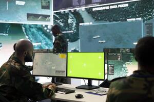 Army soldier working with an isolated pc display next to satellite footage, monitoring CCTV surveillance imagery to ensure national safety in command post. Trooper works on a mission. photo