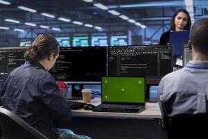 Multiracial people in data center updating systems with green screen device, protecting tech from errors. Teamworking group of engineers looking for bugs causing slow down using mockup notebook photo