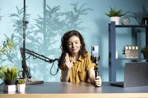 Woman greeting viewers during vlog, using smartphone on selfie stick to capture footage, chatting with online audience. Upbeat content creator saluting fans, filming with mobile phone photo