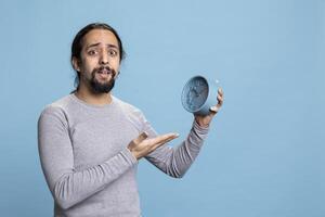 Middle eastern guy pointing at a blue wall clock to check the time in studio, wearing casual clothes and looking at the hour and minutes. Young person holding a watch to be punctual on camera. photo