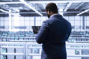 Programmer using tablet in data center to do disaster recovery planning and security enforcement. African american man in server room working to reduce equipment downtime, ensuring business continuity photo