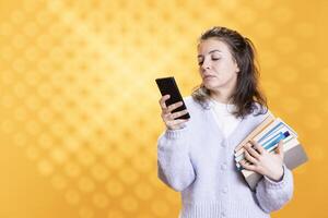 Woman reading messages on smartphone while holding stack of books, isolated over studio background. Person with pile of academic textbooks in arms texting friends on phone photo