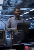 Smiling engineer pleased by recovery plan, monitoring data center energy consumption. Happy african american employee in server room making sure sensors are functioning optimally, doing disk checks photo