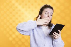 Woman yawning while reading boring book for school exam on tablet, studio background. Student feeling exhausted while researching on online textbooks using ebook reader for university assessment photo