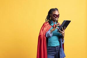 Smiling african american girl portraying superhero hiding identity, working on laptop, studio background. Jolly woman posing as hero in costume, solving tasks on notebook, covering face with mask photo