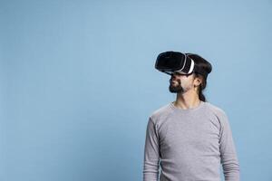 Trendy middle eastern guy using virtual reality headset on camera, using 3D artificial intelligence tech and cyberspace against blue background. Young person does gaming on a vr glasses interface. photo