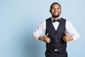 Happy confident hotel porter giving a thumbs up sign on camera, presenting his approval and trust. Satisfied smiling personnel in a tuxedo showing an okay like gesture, good vibes. photo