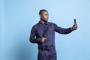 Confident bodyguard taking selfies on his smartphone with baton in studio, using a phone to take pictures against blue background. Young skilled agent has fun with photos in his uniform.