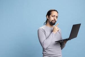 Intelligent young adult browsing on laptop symbolizing digital work and online networking, posing with device against blue background. Male freelancer surfing the web for digital communication. photo
