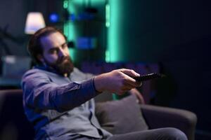Focus on remote control used by man in blurry background watching shows on TV, paying close attention to screen. Close up shot of man wearing pajamas consuming mindless entertainment photo