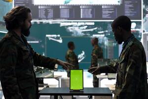 Diverse team of soldiers examine information on a green screen, working with a tablet in military base of operations. Skilled IT engineers monitoring activity in control center, high tech. photo