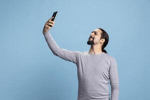 Male person discussing and laughing with his friends by conferencing, using online network on a call against blue background. Young joyful adult enjoying web discussion. photo