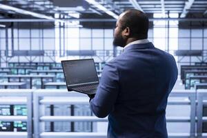 IT staff member in data center implementing backup solutions to protect against infrastructure data loss using laptop. IT professional resolving technical difficulties affecting rigs using notebook photo
