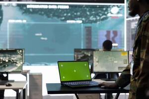 Army trooper examines green screen layout on a laptop in control center, military base of operations staff monitoring satellite world map for national defense missions. Soldier in uniform. photo