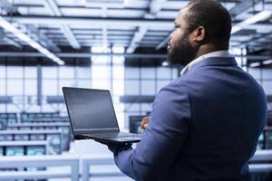 System administrator in data center making sure rackmounts are functioning optimally. African american man in server hub using laptop to monitor energy consumption across supercomputer rigs. photo