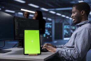 Smiling man working on green screen tablet in modern data center. Happy IT specialist doing IT infrastructure management on server racks in high tech environment using chroma key device photo