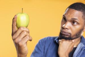 Young person holds a green apple and thinks about a dessert recipe to make with it, preparing to cook something with his ethically sourced organic produce. Pensive guy in front of camera. photo
