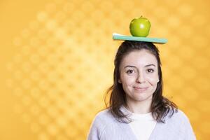 Portrait of jolly woman placing book on head and apple, acting quirky, enjoying reading hobby. Funny geek having fun wearing novel and fruit as hat, toying around, isolated over studio background photo