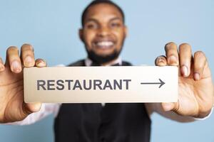 Hotel porter presenting the direction to the restaurant with a pointer, holding an indicator to guide clients to the amenities. Professional staff in tuxedo using a wall sign. Close up. photo