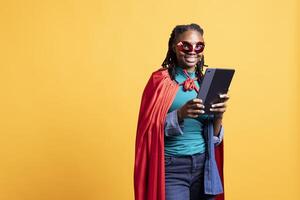 Portrait of smiling african american girl portraying superhero hiding identity, using tablet, studio background. Jolly woman posing as hero in costume, using digital device, covering face with mask photo