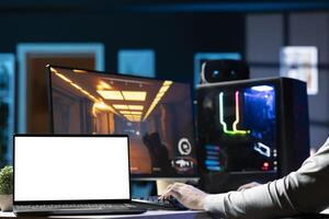 Man sitting on gaming chair using mockup laptop, relaxing by playing videogame on PC at computer desk late at night. Isolated screen notebook next to gamer enjoying game on computer photo