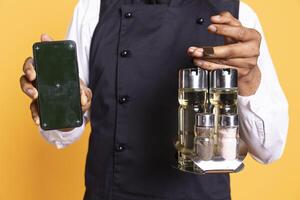 Young butler carrying a cruet set and phone with copy space display, posing with apron against yellow background. Professional caterer working in service oriented industry. photo