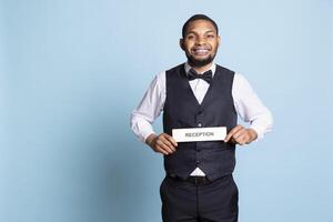 Hotel concierge staff showing the reception indicator at front desk, greeting guests at five star holiday resort in studio. Skilled professional bellboy holds sign against blue background. photo