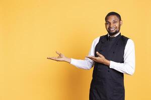 African american waiter points at something to his left or right, indicating direction to the sides in studio. Customer focused employee wears uniform and apron against background. photo