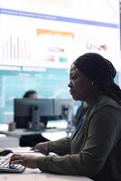 Marketing specialist studying infographics on computer in a monitoring room with big screen, check customer satisfaction from annual data reports. Employee review performance metrics. photo