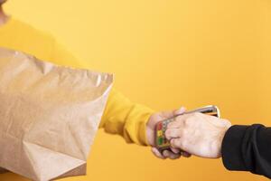 Client paying with credit card to part time delivery worker holding lunch meal order. Close up shot of man purchasing order in paper bag from courier, swiping card on POS device, studio background photo