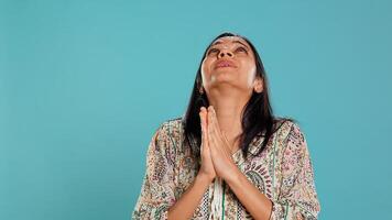 Indian woman putting hands together in begging gesture, making wish, isolated over studio background. Faithful person looking upwards to sky, asking for something, praying and hoping, camera B photo