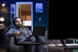 Man sitting in neon lit apartment on couch working on laptop while picking TV channel to watch as distraction. Remote worker solving company tasks on notebook and browsing through shows using remote photo