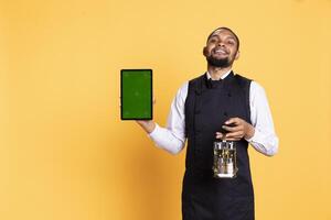 Caterer with apron and cruet set shows green screen on tablet, working in catering customer focused industry. Professional butler in uniform holds a gadget with isolated display. photo