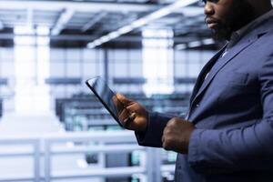 Data researcher using tablet in high tech data center, ensuring cybersecurity and efficient network management. Employee in server hub optimizing data center gear with device photo