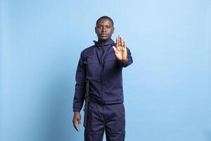 African american security agent raising his palm and saying no, showing a stop sign against blue background. Professional guarding agent does not let people pass, authority and confidence. photo
