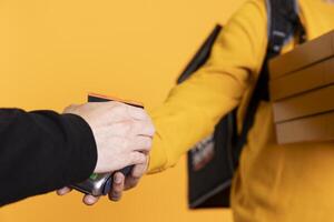Customer paying with credit card to part time delivery worker holding takeaway order. Close up shot of customer buying meal from courier, swiping card on POS device, studio background photo