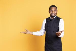 Smiling friendly butler pointing direction to left or right sides against yellow background, creating an advertisement for the catering industry. Five star restaurant employee. photo